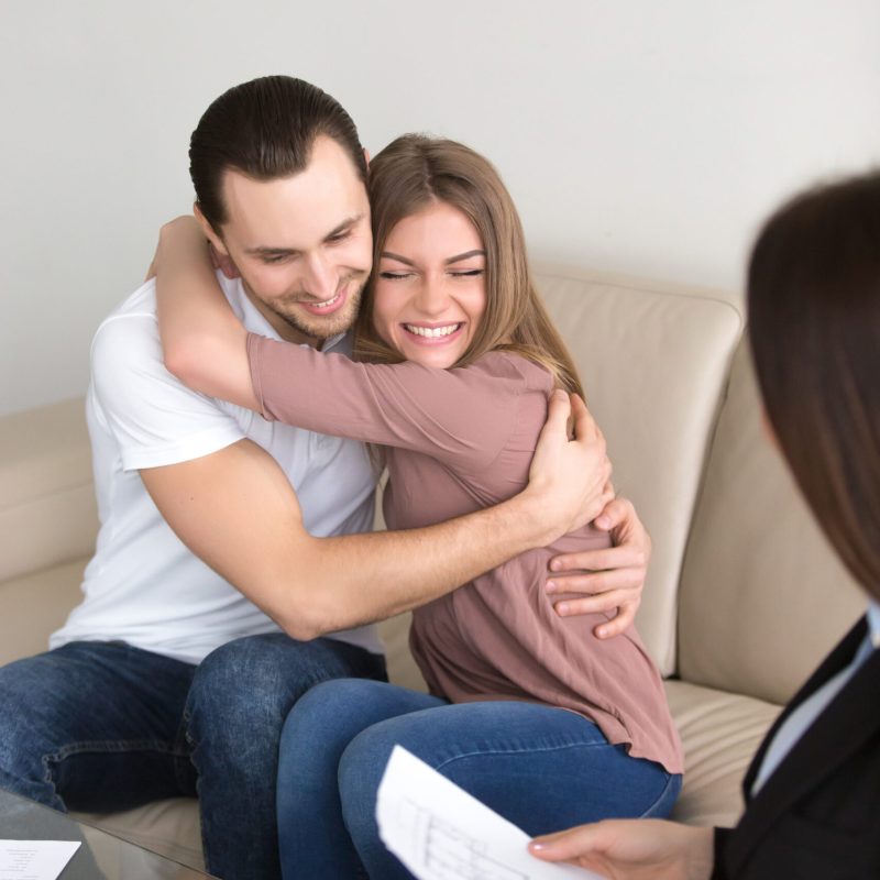 Happy young couple ready to buy apartment excited about first investment in property, cheerful woman likes new home, beautiful smiling wife hugging her husband sitting on couch with real estate agent